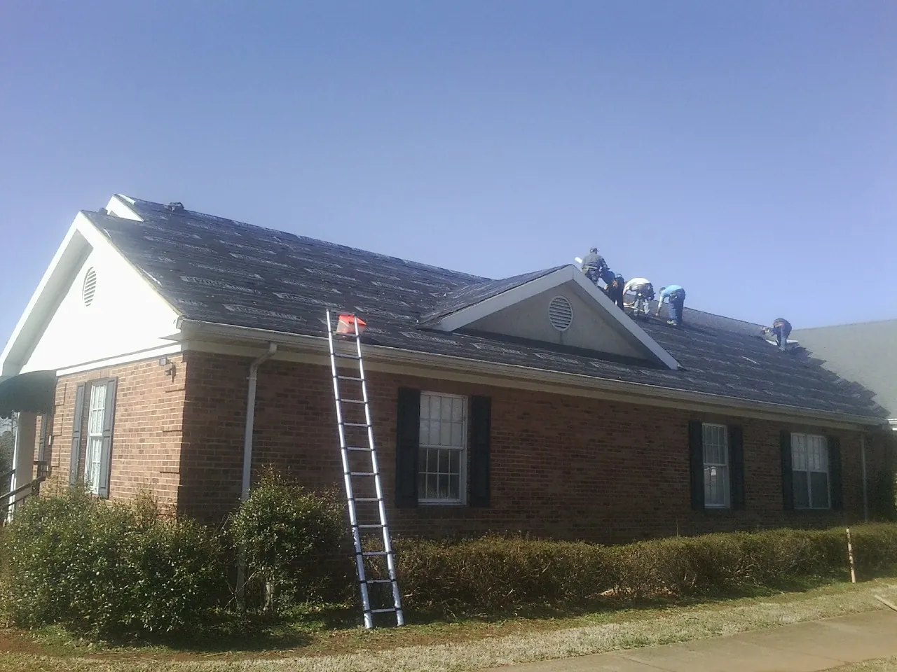 Crew tearing shingles off a brick ranch roof with a ladder set — Metal Roof Repair work by our crew in Partlow