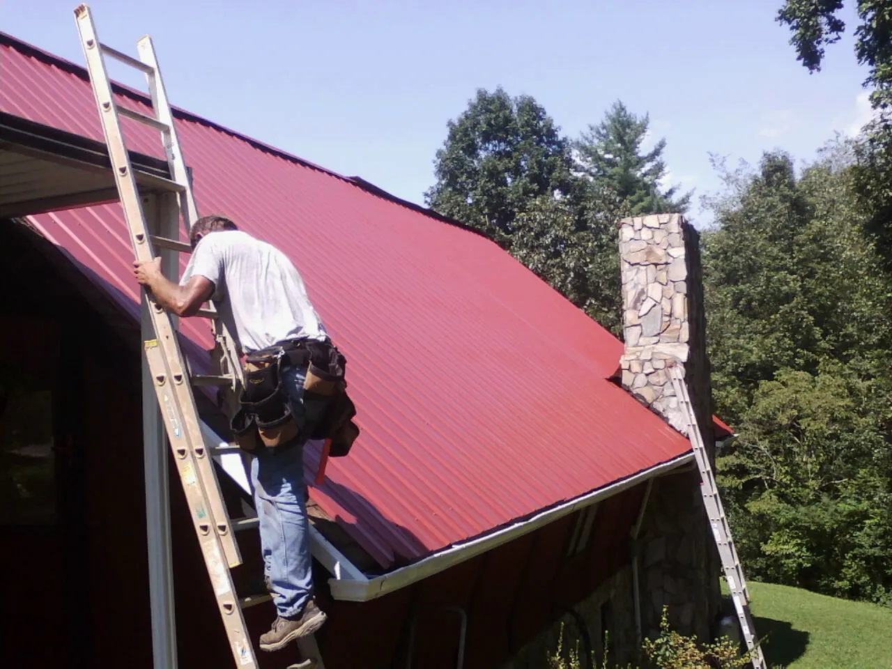 Roofer on a ladder beside a red metal roof and stone chimney — Soffit & Fascia Repair by Roofing Brothers in Partlow